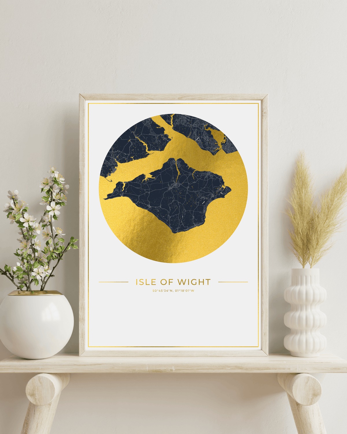 A framed art print of the Isle of Wight, England, featuring a gold foil map silhouette on a white background with a light wood frame. The print is styled on a light wooden shelf next to white ceramic vases containing green and beige dried botanicals against a neutral off-white wall.
