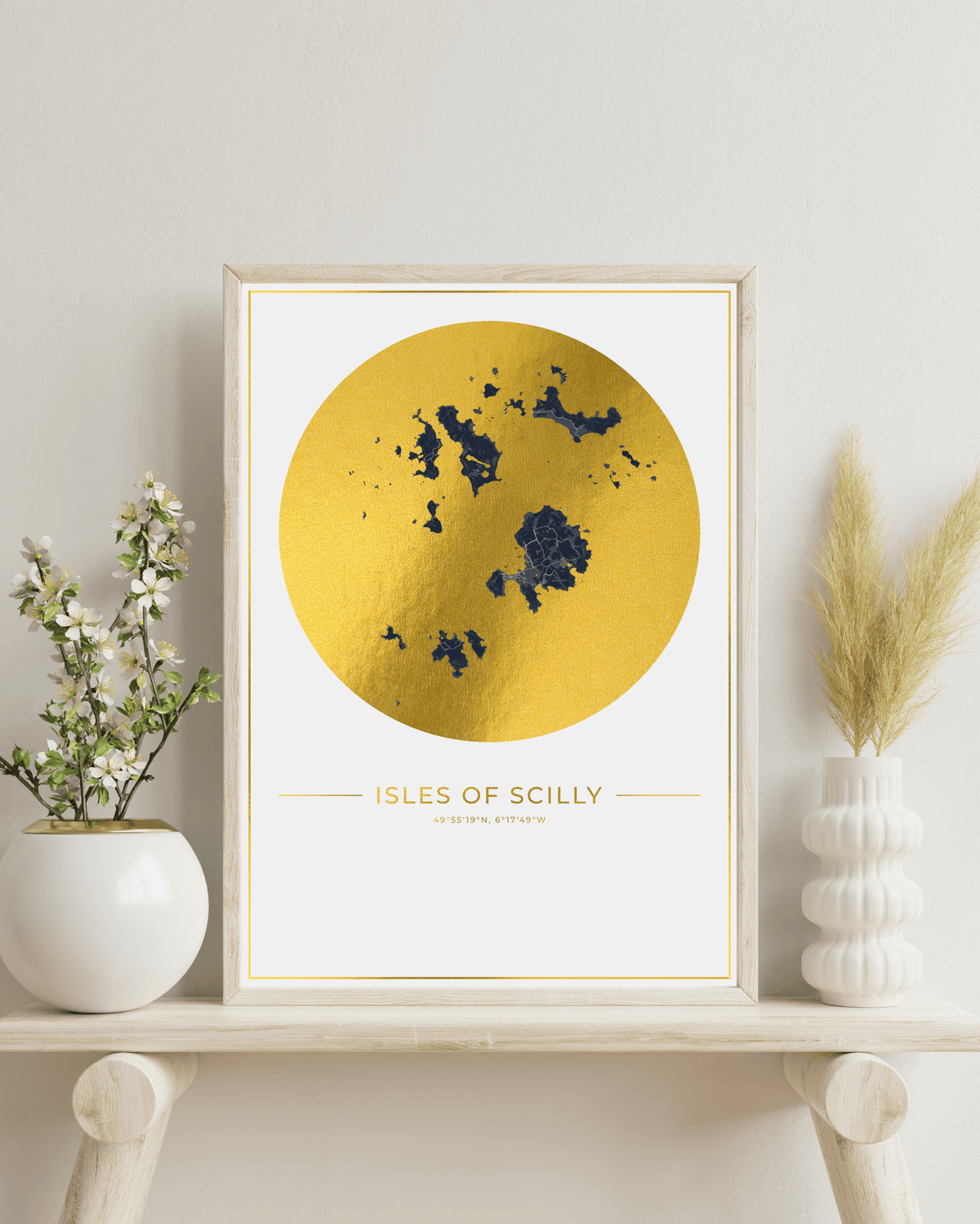 A framed art print featuring a textured gold circle with a dark blue map of the Isles of Scilly and the text 'ISLES OF SCILLY' on a white background, styled on a light wood shelf with decorative vases.