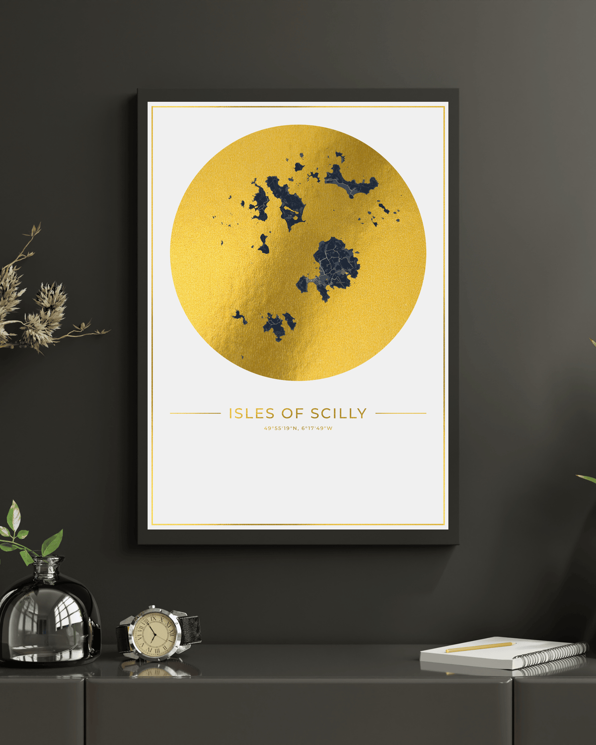 A framed wall art print featuring a large gold circle with a dark blue map of the Isles of Scilly and the text 'ISLES OF SCILLY' below, displayed on a dark gray wall above a console table with decorative objects.