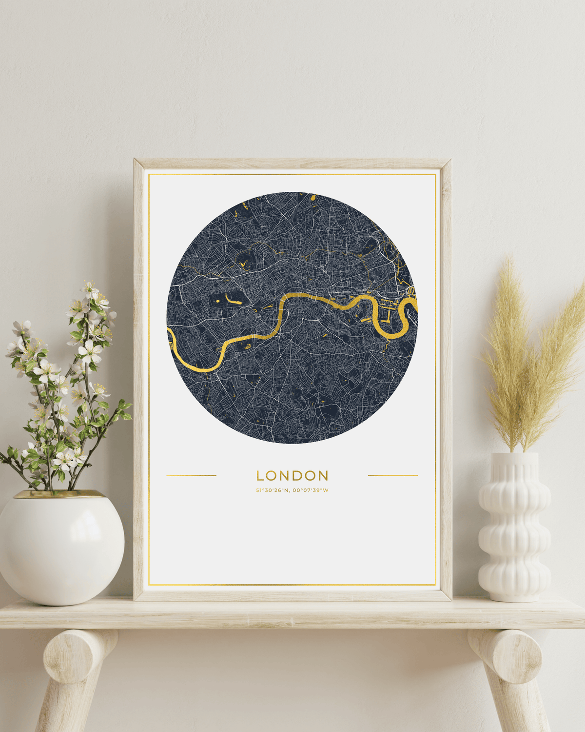 A framed London city map art print with a dark navy blue circular map design highlighted by a yellow River Thames curve, displayed on a light wood shelf with decorative vases and dried grass.