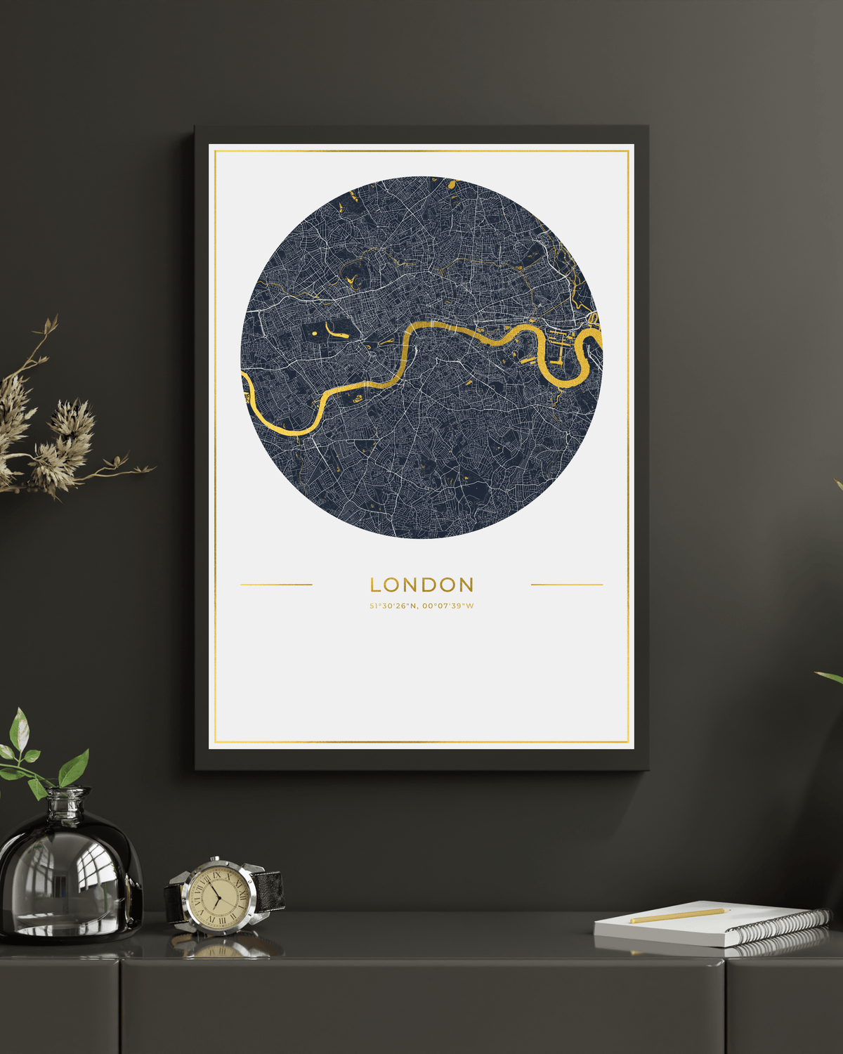 A framed London city map art print with a dark navy circular map design, gold River Thames line, and gold text on a white background, hanging on a dark gray wall above a console table with decorative objects.