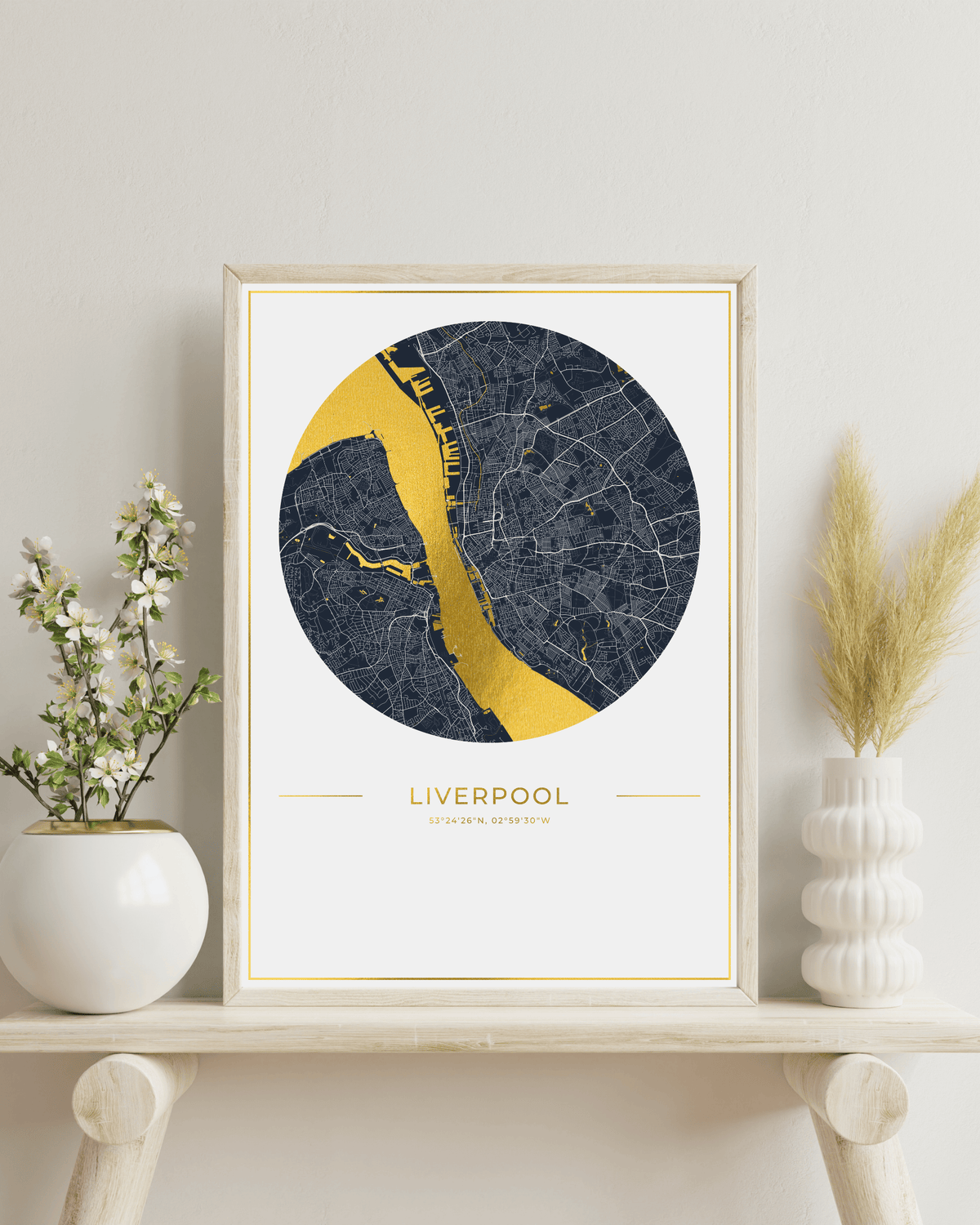 A framed circular map art print of Liverpool, England, featuring a dark navy street network and a bright gold river, displayed on a light wood shelf with decorative vases.