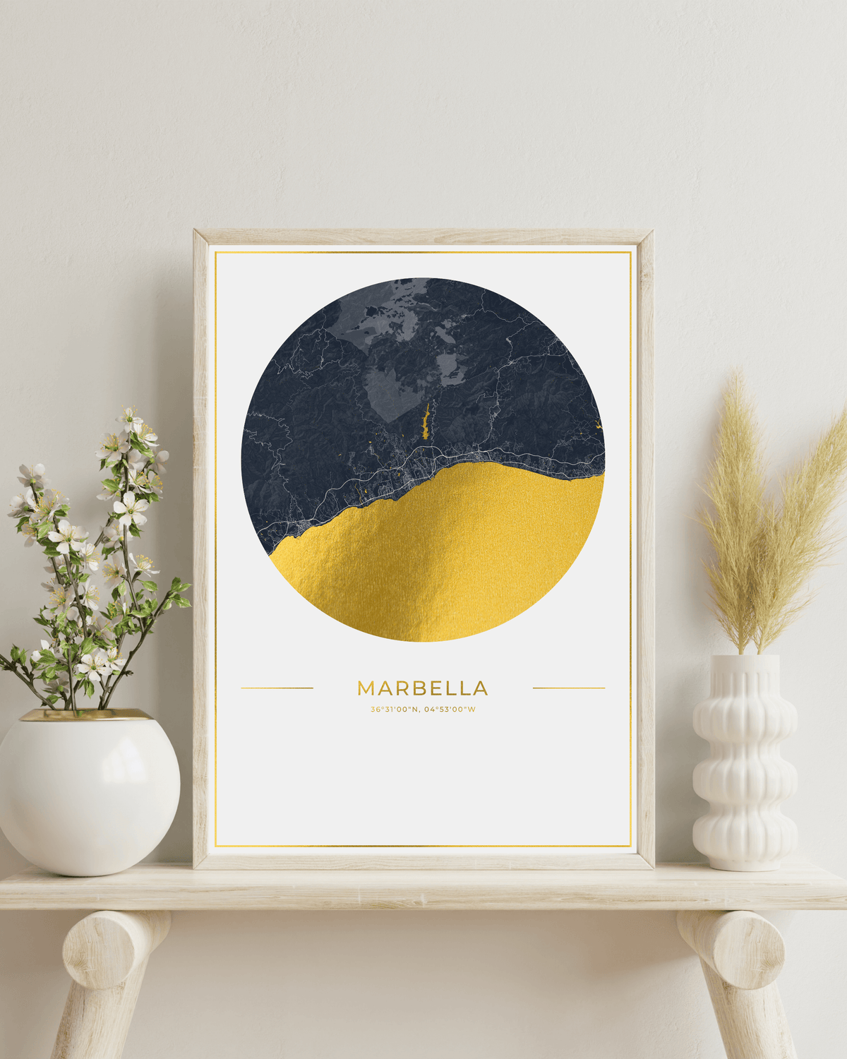 A framed art print featuring a circular design with a dark navy marble top half and a textured gold bottom half, displayed on a light wood shelf with decorative plants and vases against a neutral wall.