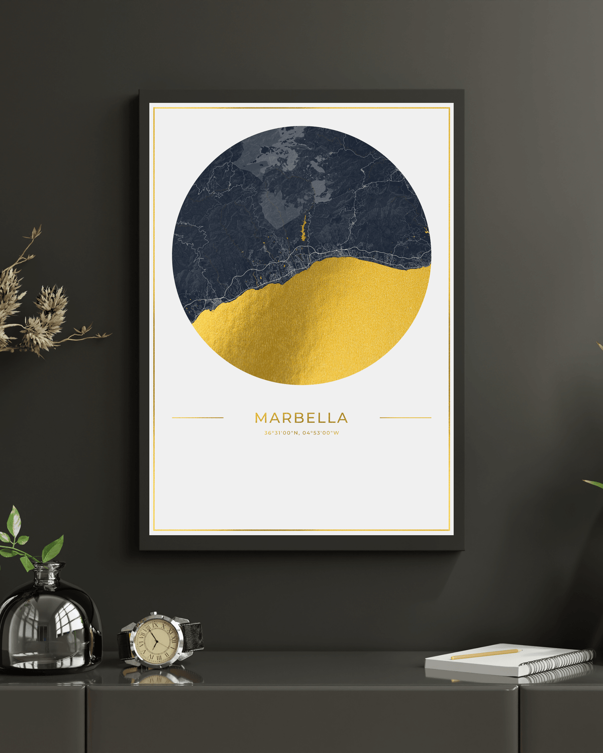 A framed art print featuring a circular map of Marbella with a dark navy blue marble texture and gold foil accents, displayed on a dark gray wall above a console table with decorative items.