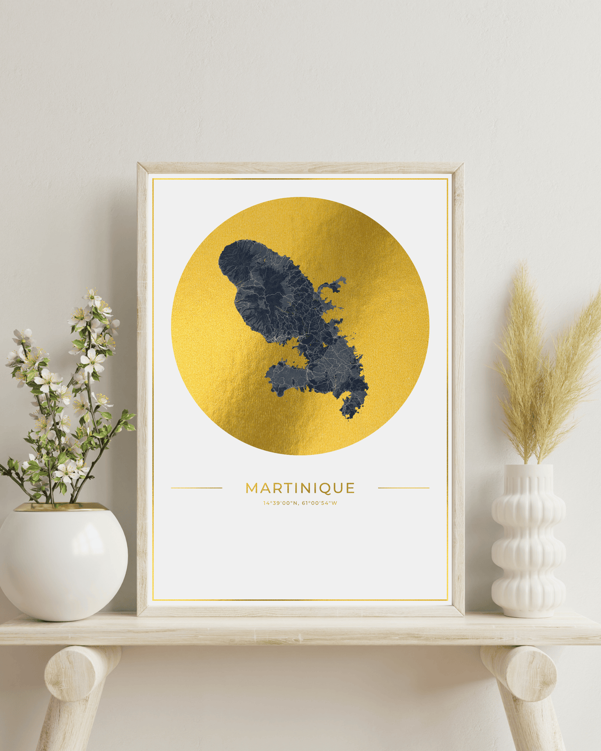 Framed wall art featuring a dark blue map silhouette of Martinique over a large metallic gold circle on a white background, hung on a LIGHT WOOD  table with decorative objects.