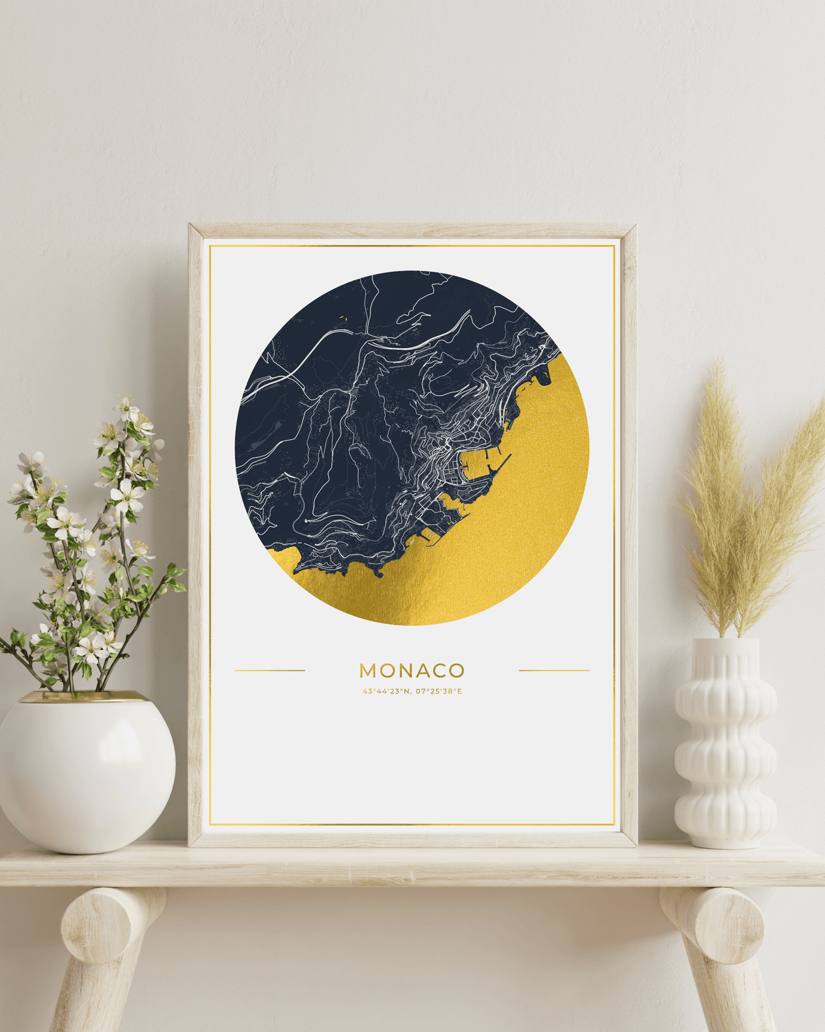 A framed art print of a stylized map of Monaco, featuring a navy blue and gold circular design with white city outlines, displayed on a wooden shelf next to decorative vases.