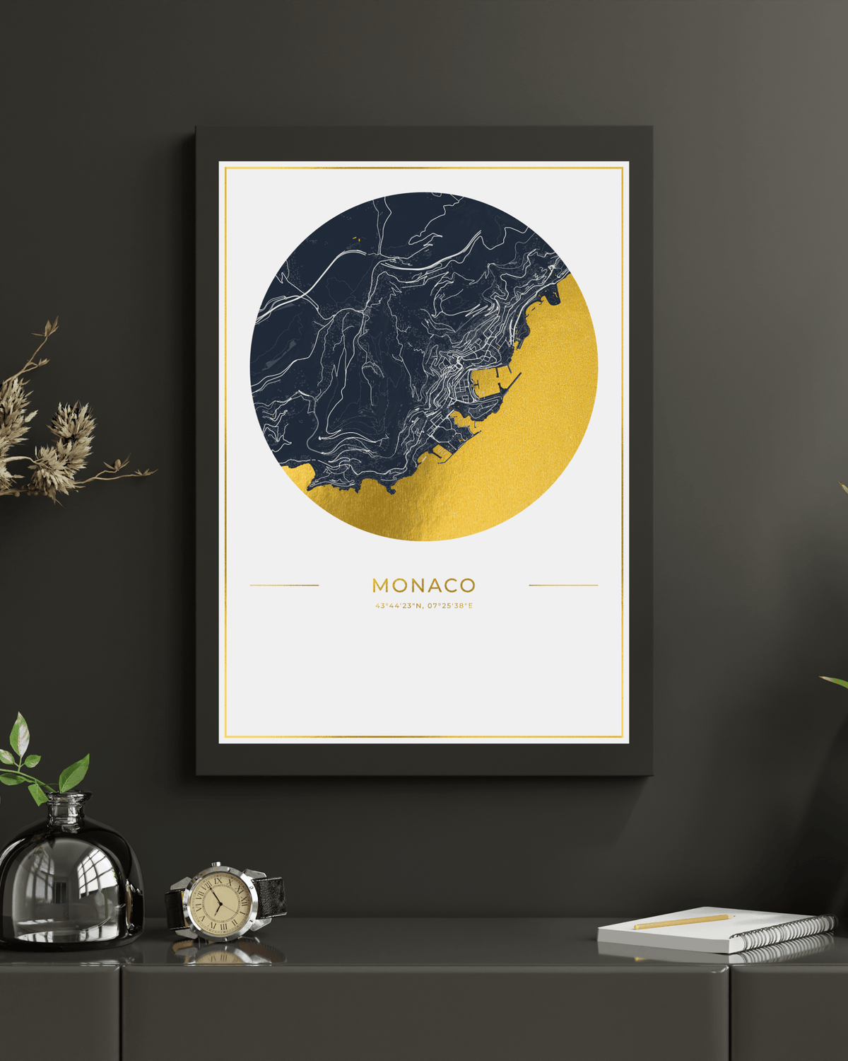A framed art print of Monaco featuring a circular map design in navy blue and gold, displayed on a dark gray wall above a console table with decorative items.