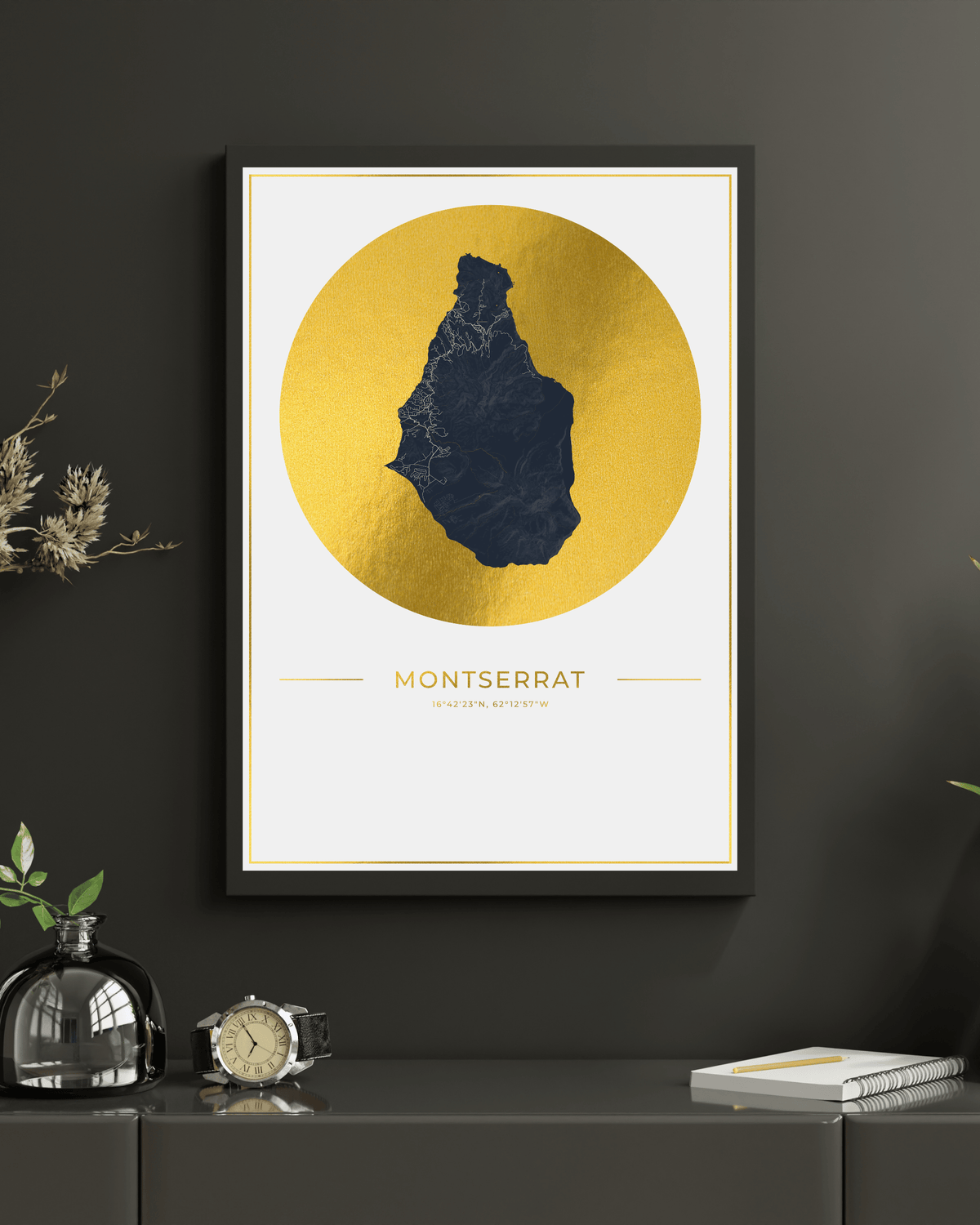 A framed art print of Montserrat, featuring a dark blue textured silhouette of the island on a large gold circular background, mounted on a dark gray wall above a console table with decorative items.