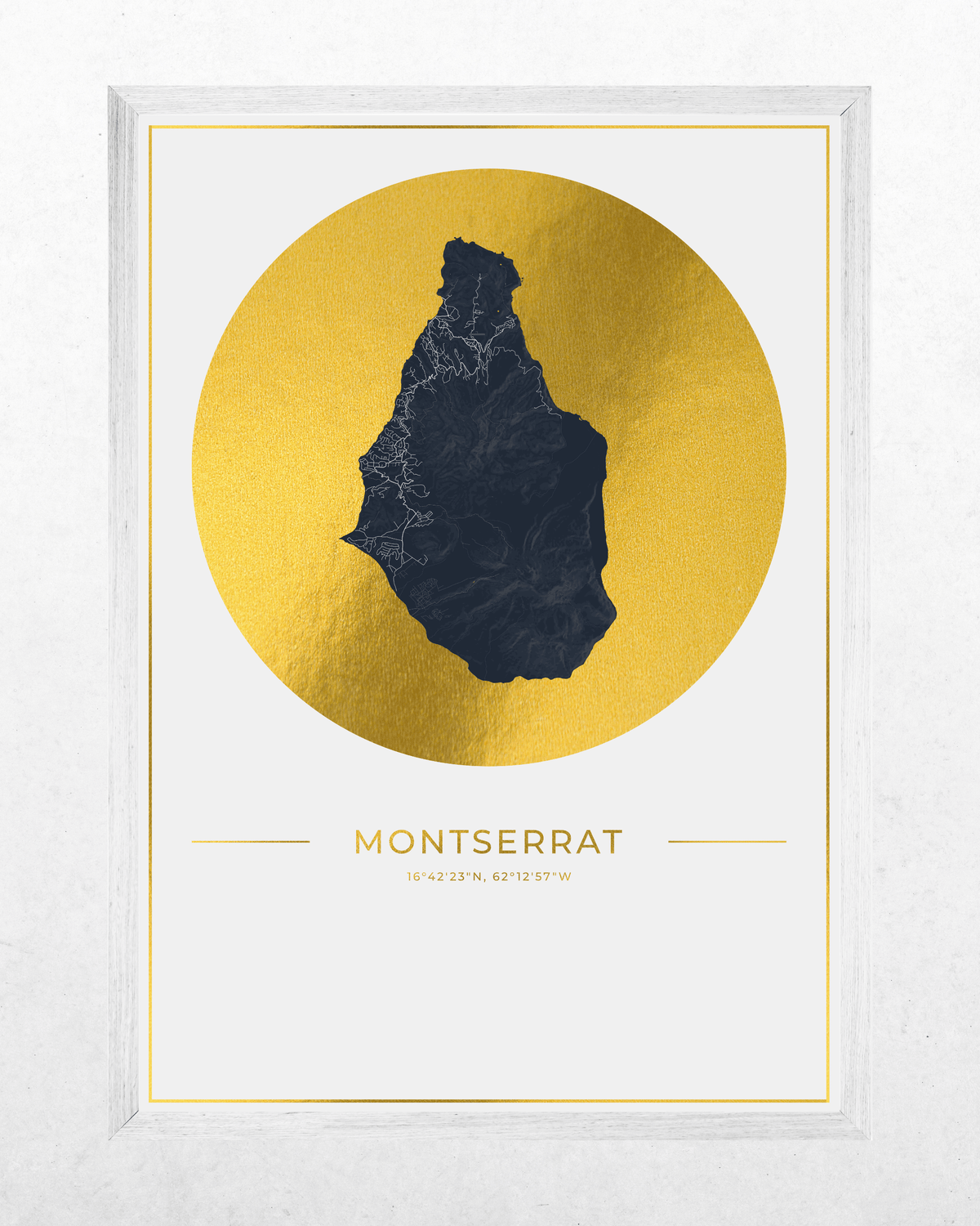 Framed wall art print featuring a dark navy silhouette map of Montserrat centered inside a large metallic gold circle, with the word 'MONTSERRAT' and coordinates printed below on a white background. The frame is light gray and shown against a textured white wall.