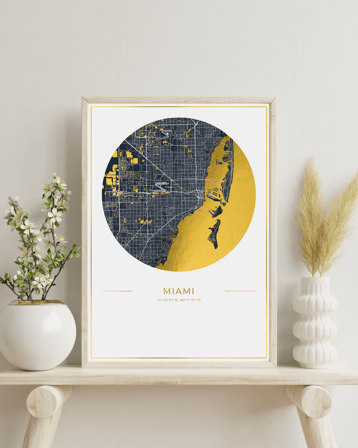 A framed art print featuring a stylized map of Miami, Florida, with a navy blue street grid and a large gold coastal shape, displayed on a light wood shelf with decorative vases against a light gray wall.
