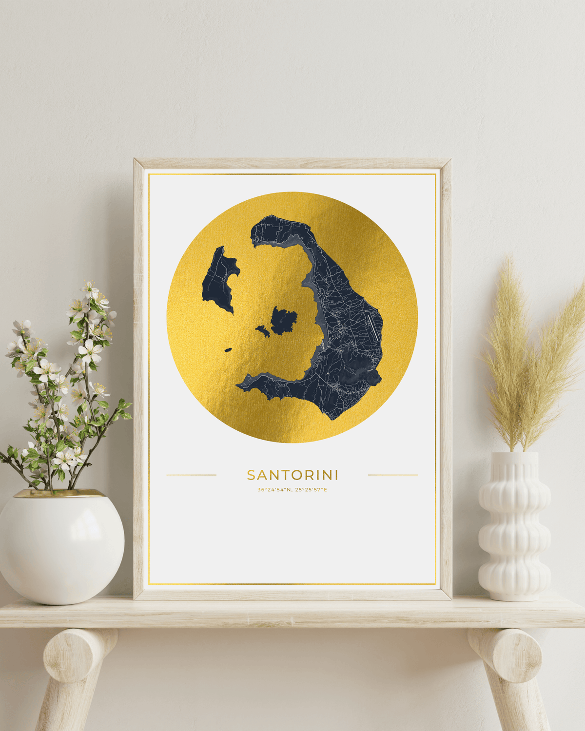 A framed art print featuring a gold map of Santorini, Greece, styled on a light wood shelf with decorative white vases and dried grass against a neutral wall.