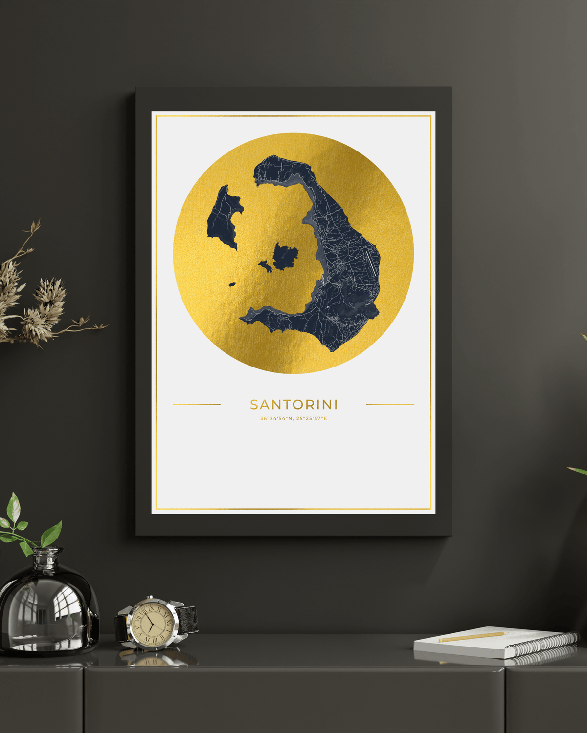 A framed art print featuring a gold map of the island of Santorini on a white background, hanging on a dark gray wall above a console table with decorative objects.
