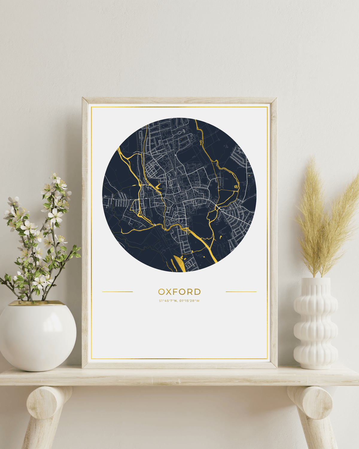 A framed art print featuring a circular street map of Oxford in navy blue and gold, styled on a light wood shelf with white ceramic vases and dried pampas grass against a neutral wall.