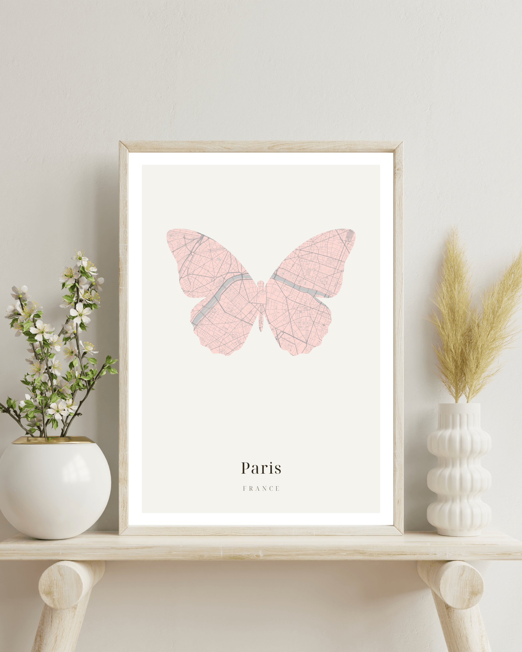 A framed butterfly art print featuring a pink butterfly map design with the text 'Paris FRANCE' at the bottom, displayed on a light wood shelf with decorative plants and vases against a neutral wall.