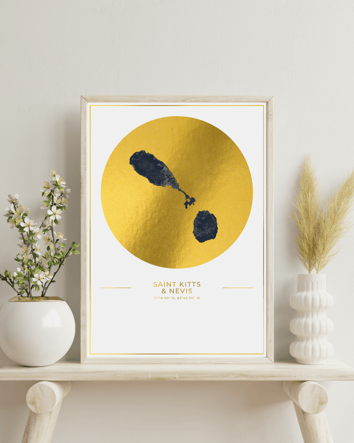 A framed art print featuring a textured gold circle with a map of Saint Kitts & Nevis on a white background, styled on a wooden shelf with potted plants and dried grass in white ceramic vases.