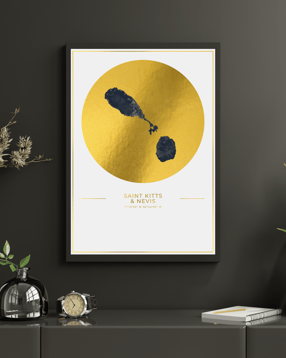 A framed minimalist art print featuring a textured gold circle with a map of Saint Kitts & Nevis on a white background, hung on a dark gray wall above a console table with decorative objects.