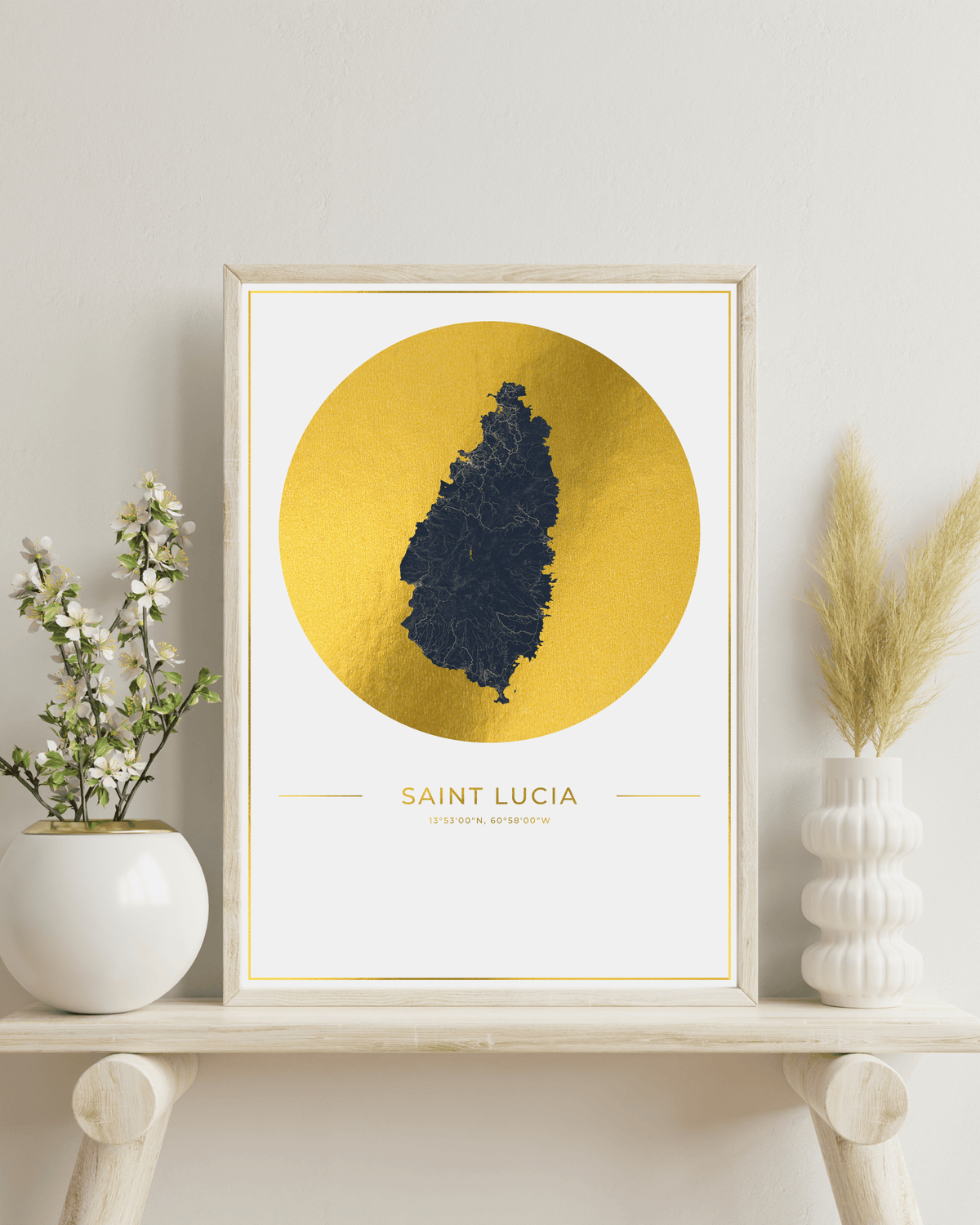 A framed art print featuring a dark blue silhouette map of Saint Lucia on a textured gold circular background, styled on a light wood shelf with white ceramic vases and dried pampas grass against a neutral wall.