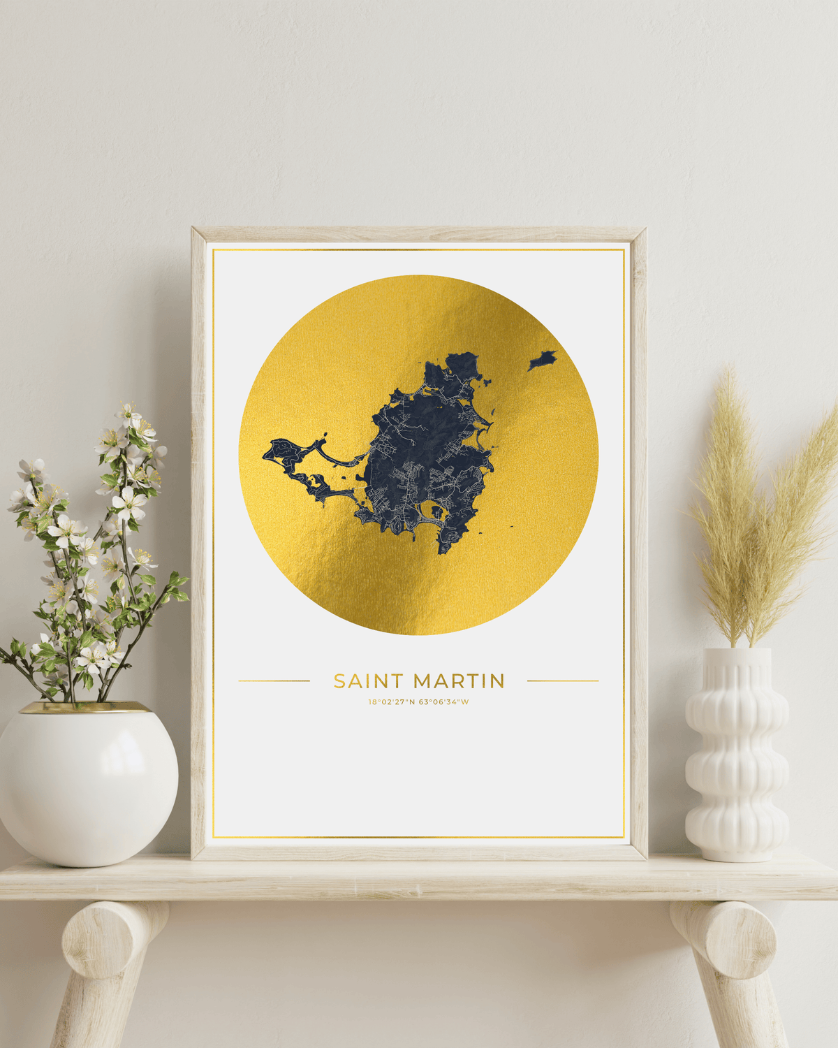 A framed art print featuring a gold map silhouette of Saint Martin, France, displayed on a light wood shelf with decorative white vases and dried grass against a neutral wall.