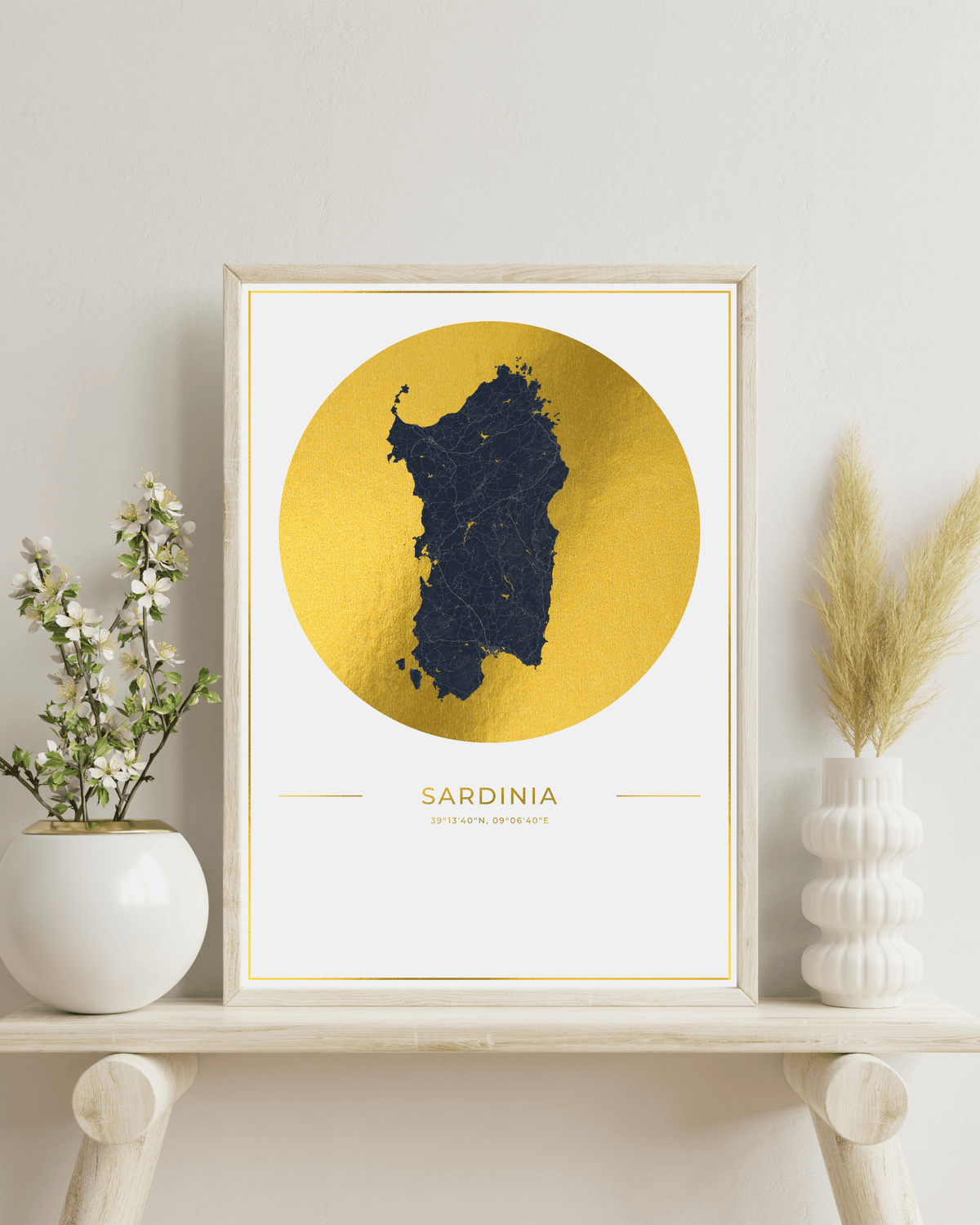 A framed art print of Sardinia, Italy, featuring a dark blue textured map silhouette inside a large gold circle on a white background. The print is housed in a light wood frame and styled on a light wooden shelf with white ceramic vases containing green and beige dried botanicals against a neutral off-white wall.