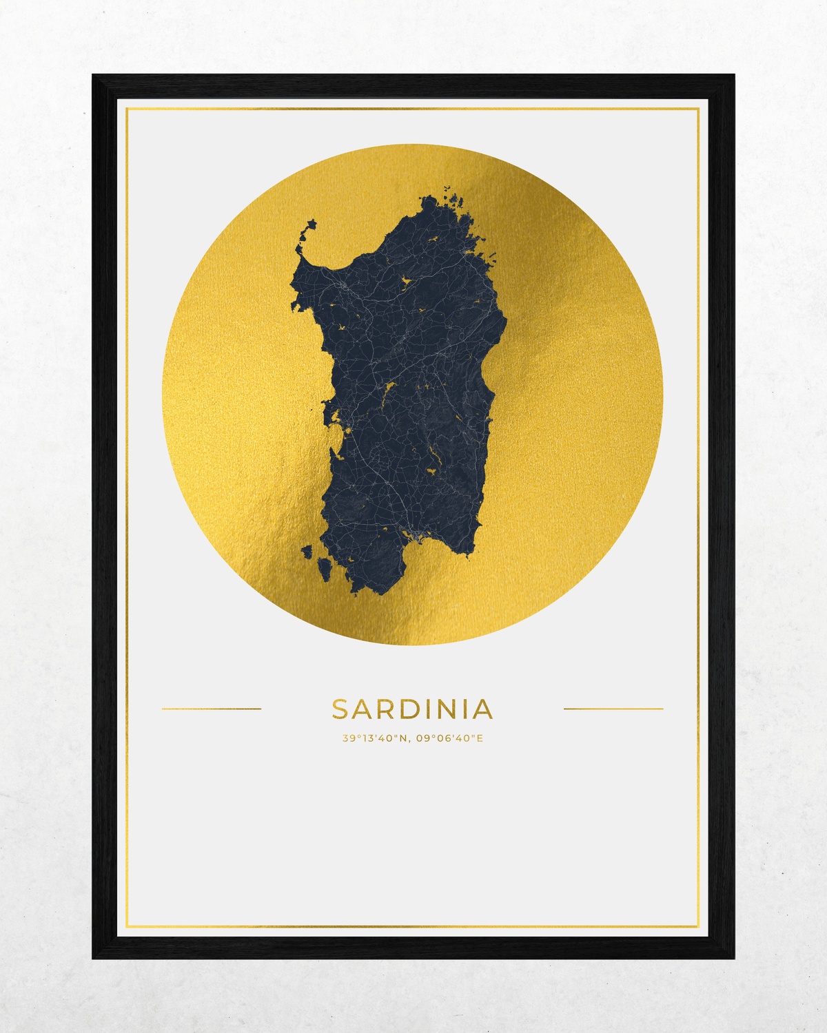 A framed art print featuring a dark navy blue map silhouette of Sardinia centered inside a large textured gold circle on a white background, with the word SARDINIA and coordinates printed below. The print is enclosed in a thin white mat and a black frame, shown against a light gray wall.