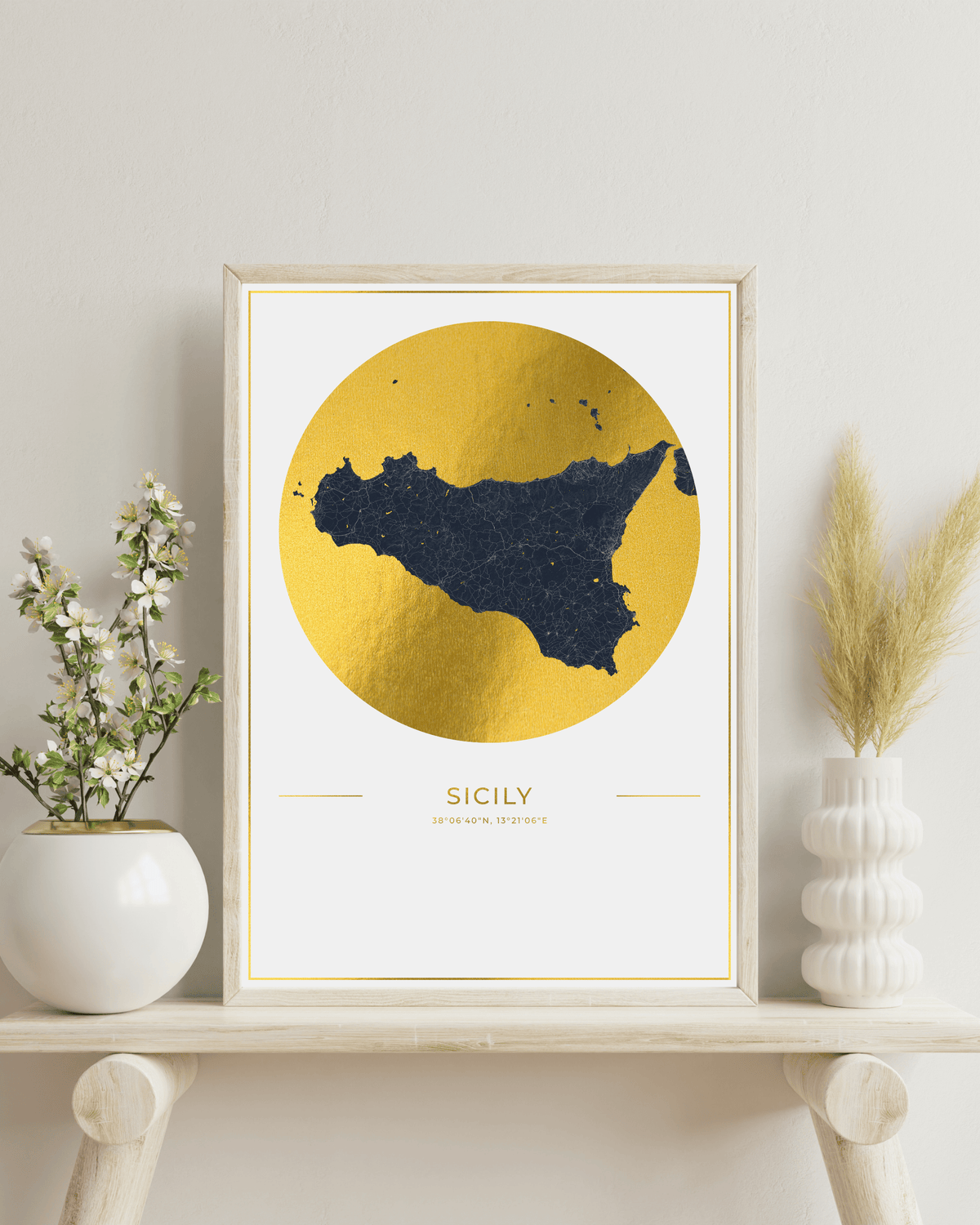 A framed art print featuring a dark navy blue silhouette map of Sicily inside a large textured gold circle, displayed on a light wood shelf next to white ceramic vases with dried grass and small white flowers.