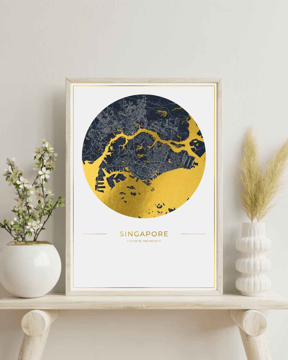 A framed Singapore city map art print with a gold and navy blue design, styled on a light wood shelf with white ceramic vases and dried pampas grass against a neutral wall.