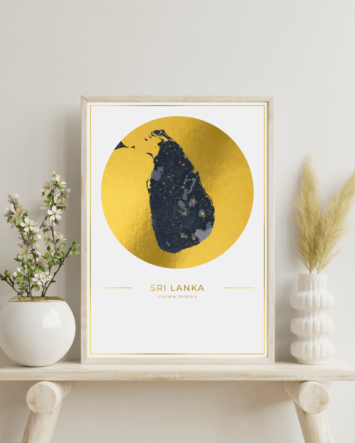 A framed art print of Sri Lanka featuring a textured gold circular background with a dark blue silhouette of the island, displayed on a light wood shelf with decorative plants.