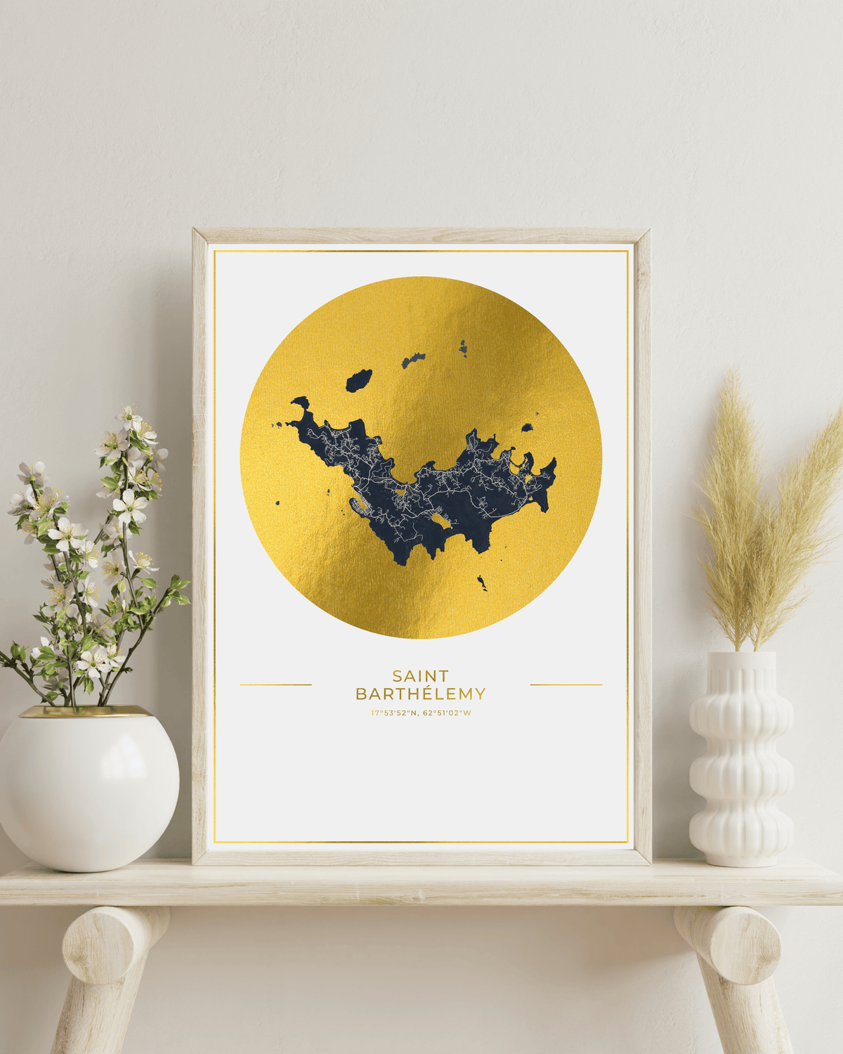 A framed art print featuring a gold map of Saint Barthelemy on a white background, styled on a light wood shelf with a white vase of flowers on the left and a white vase of dried grass on the right.