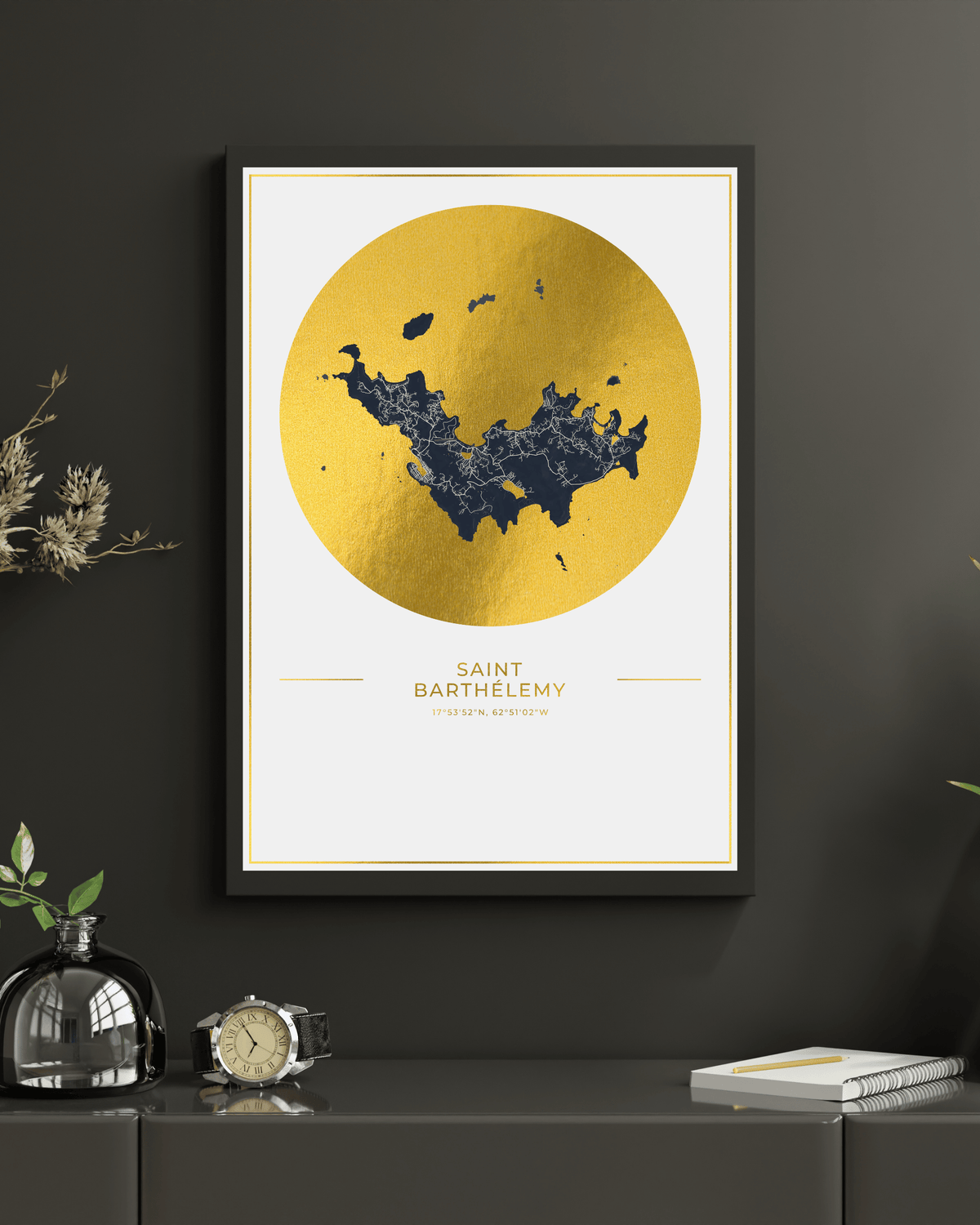A framed minimalist map print of Saint Barthelémy, featuring a textured gold circle with a dark blue map silhouette, displayed on a dark gray wall above a console table with decorative items.