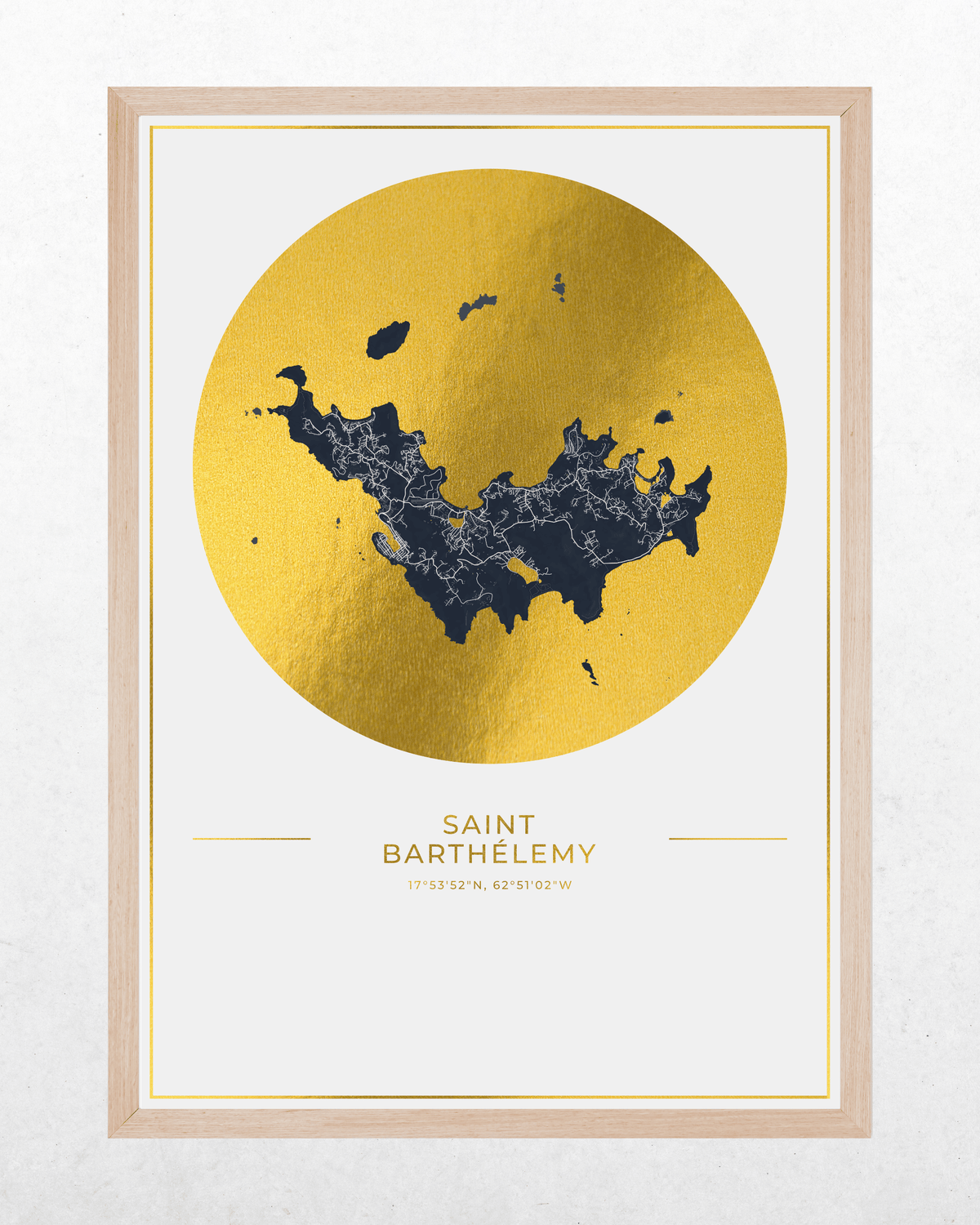 Framed wall art print featuring a gold circular map design with a dark blue Saint Barthelmy coastline, plus the city name and coordinates printed below, shown in a light wood frame against a white wall.