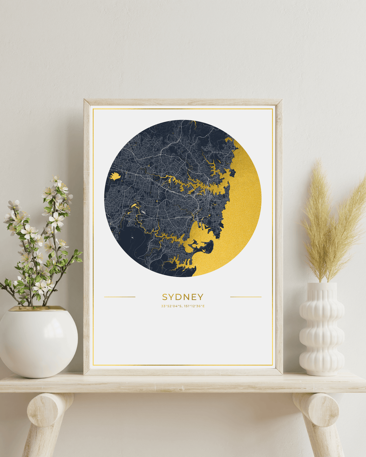 A framed Sydney city map art print with a navy and gold circular design, displayed on a light wood shelf next to white ceramic vases with dried grass and small white flowers.