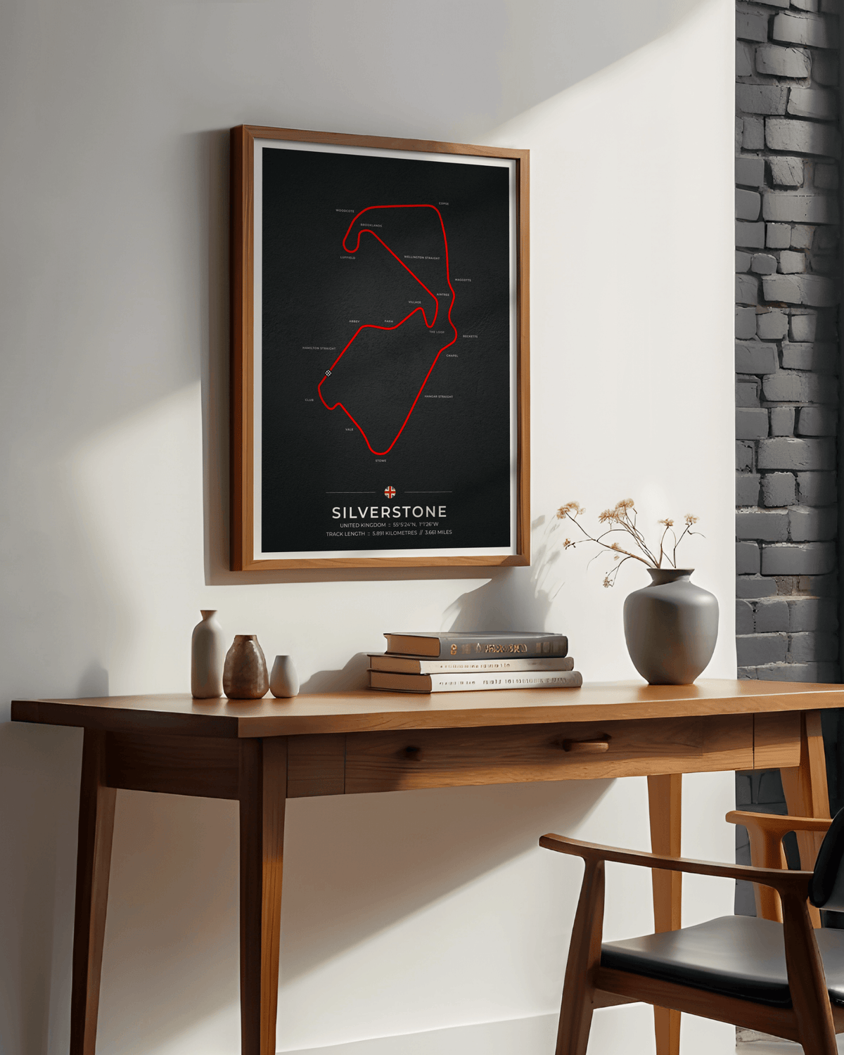 A framed poster featuring a red outline map of the Silverstone race track on a black background, hanging on a white wall above a wooden desk with books and vases.