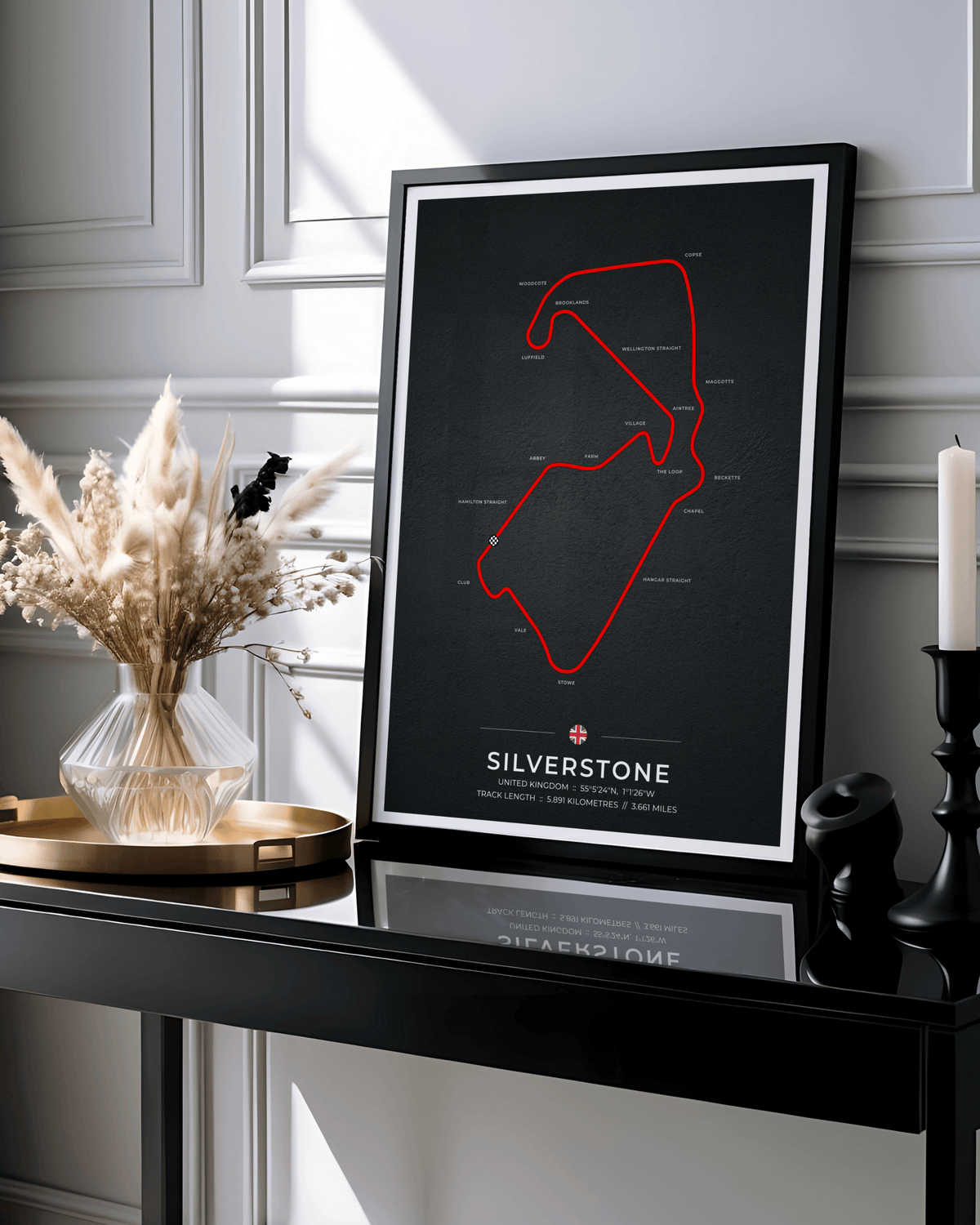 A framed poster featuring a red circuit track diagram on a black background with the word 'SILVERSTONE' at the bottom, styled on a black console table beside a vase of dried grasses and a candle in a real interior setting.