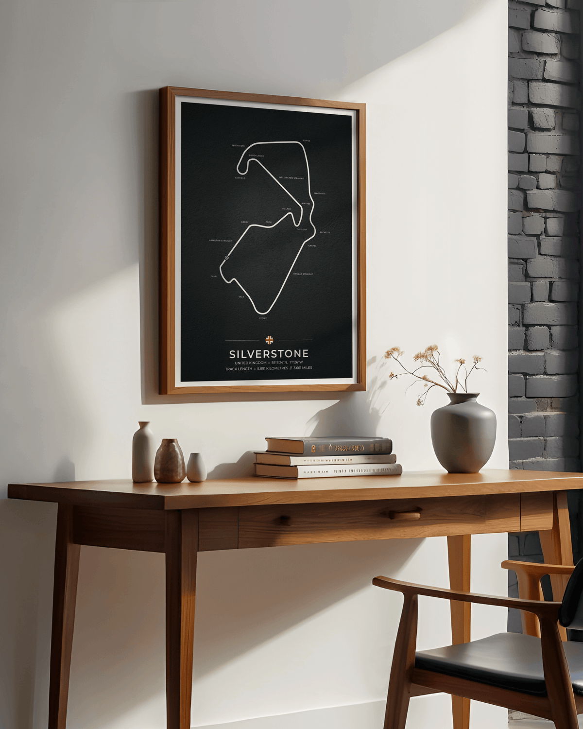 A framed black poster with a white line drawing of the the Silverstone circuit hangs on a white wall above a wooden console table in a styled interior with books, vases, and a chair.