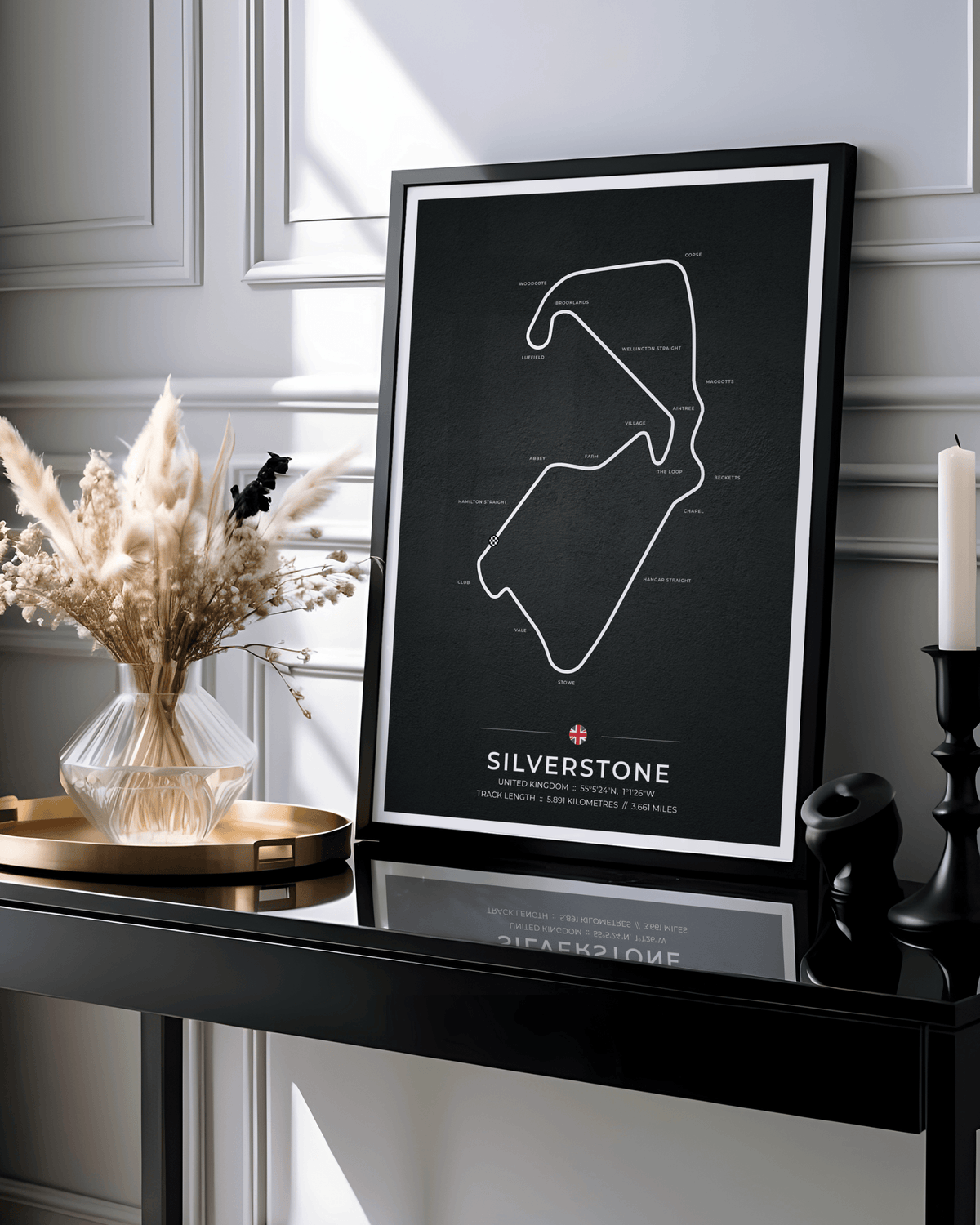 A framed black-and-white Silverstone motorsport track poster displayed on a glossy black console table in a styled interior setting with dried grasses, a candle, and decorative objects.