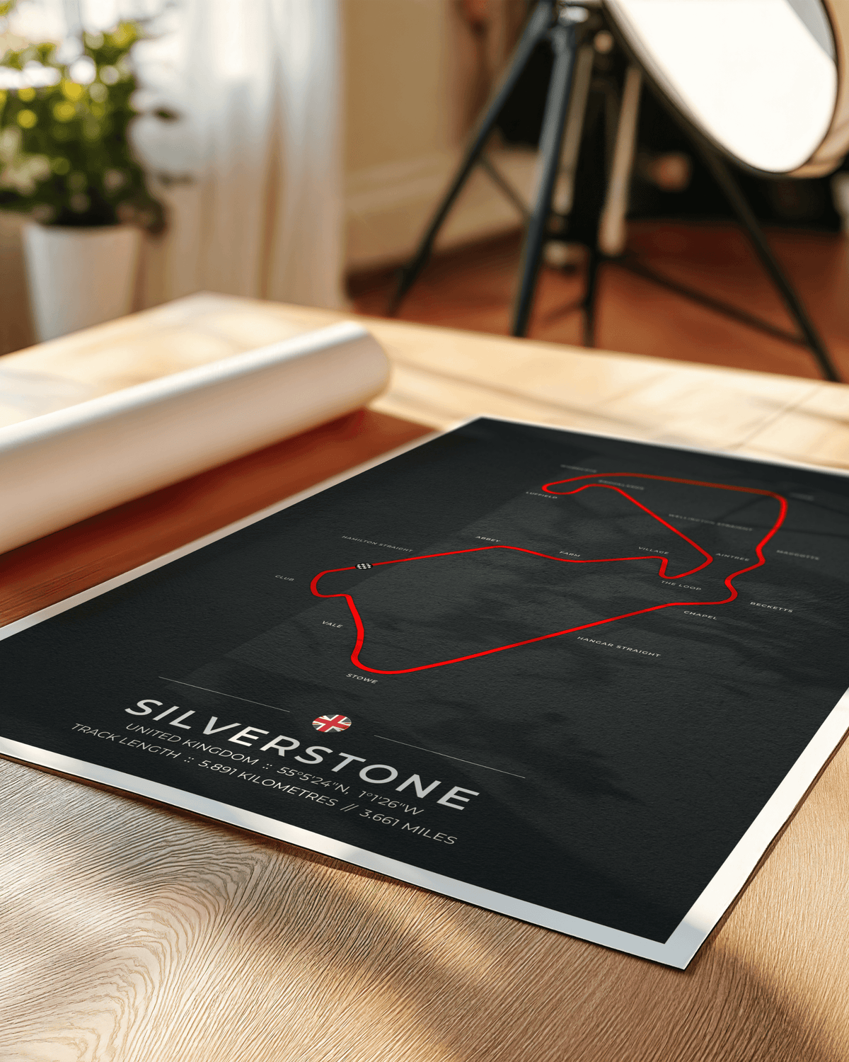 A framed Silverstone race track poster lies on a light wood tabletop in a real indoor setting, with a rolled sheet and soft-focus studio lights in the background. The poster has a dark charcoal background with a red track outline and white 'SILVERSTONE' text.