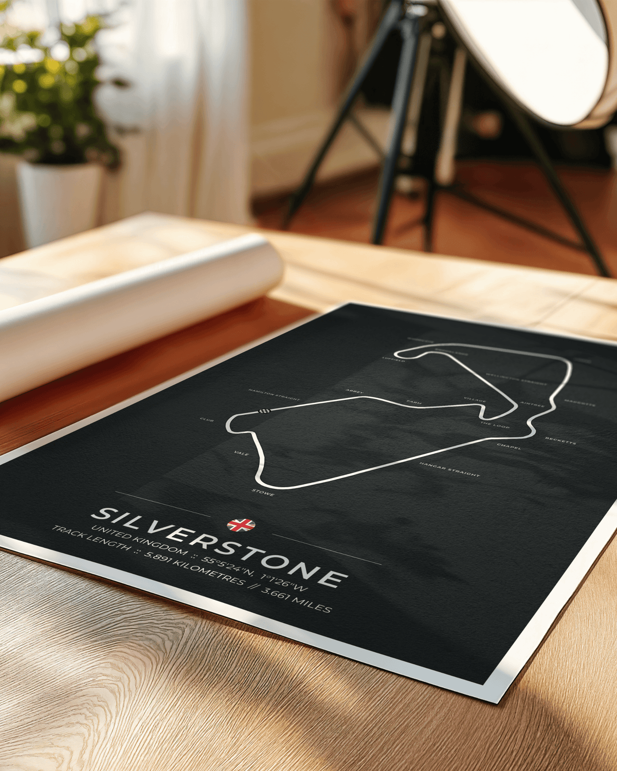 A framed poster print of the Silverstone race track layout, displayed on a light wood tabletop with a rolled print and studio lighting equipment in the background.