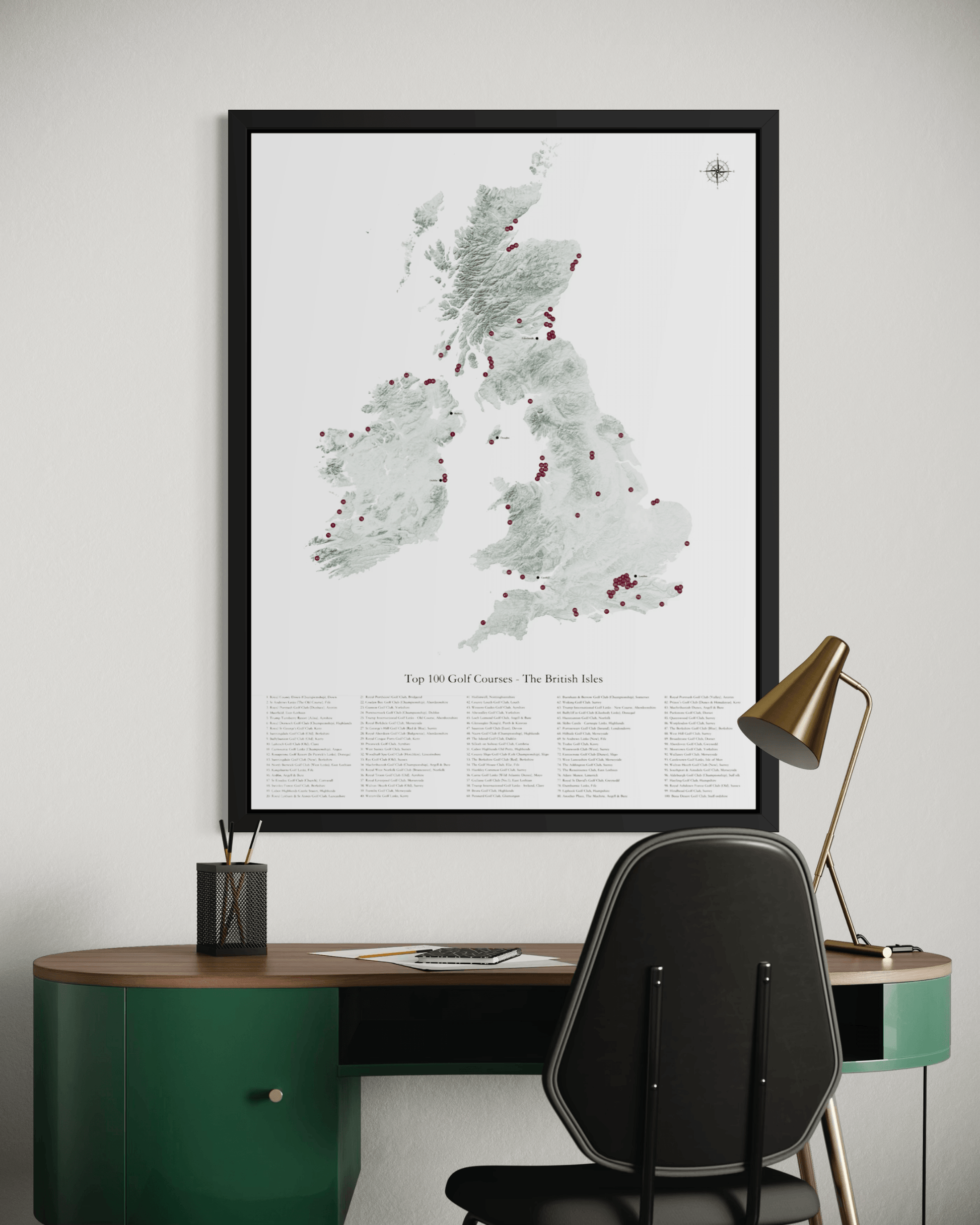 A framed vertical map print of the British Isles featuring top 100 golf courses, styled on a light gray wall above a green desk with a black chair and a gold desk lamp.