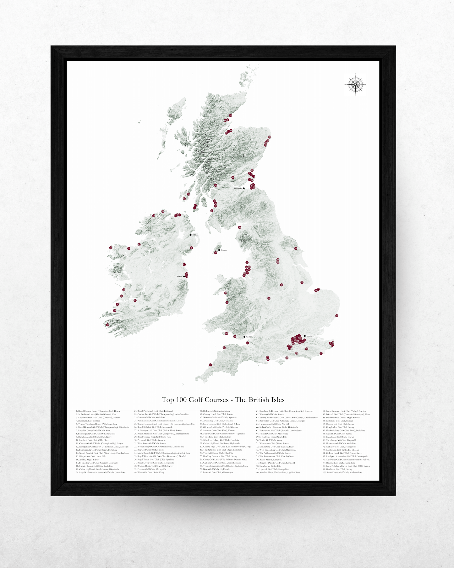 A framed vertical map print of the British Isles showing the top 100 golf courses, with a black frame on a light textured wall.