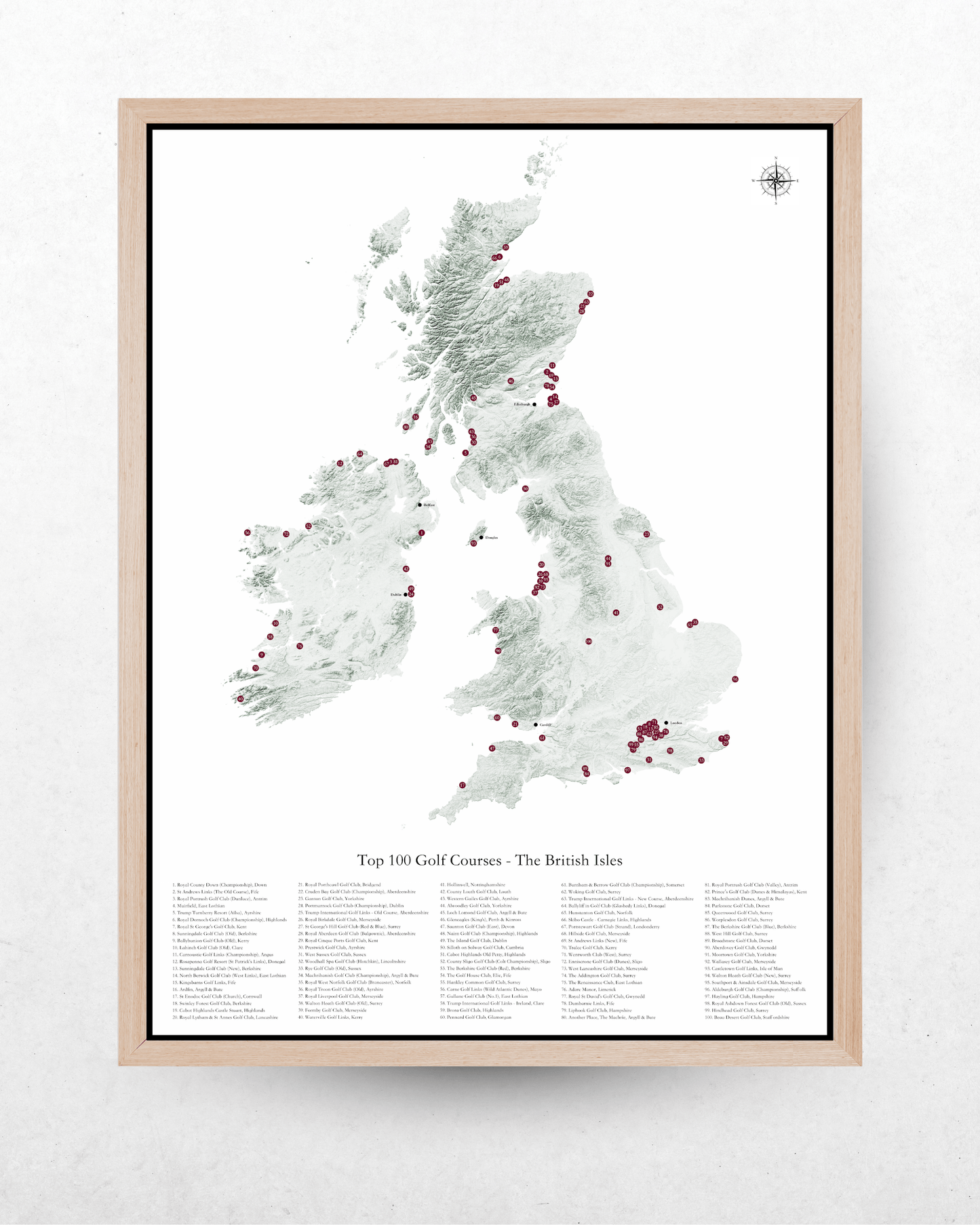 A framed map print of the British Isles highlighting the top 100 golf courses, featuring a light wood frame against a textured white wall.