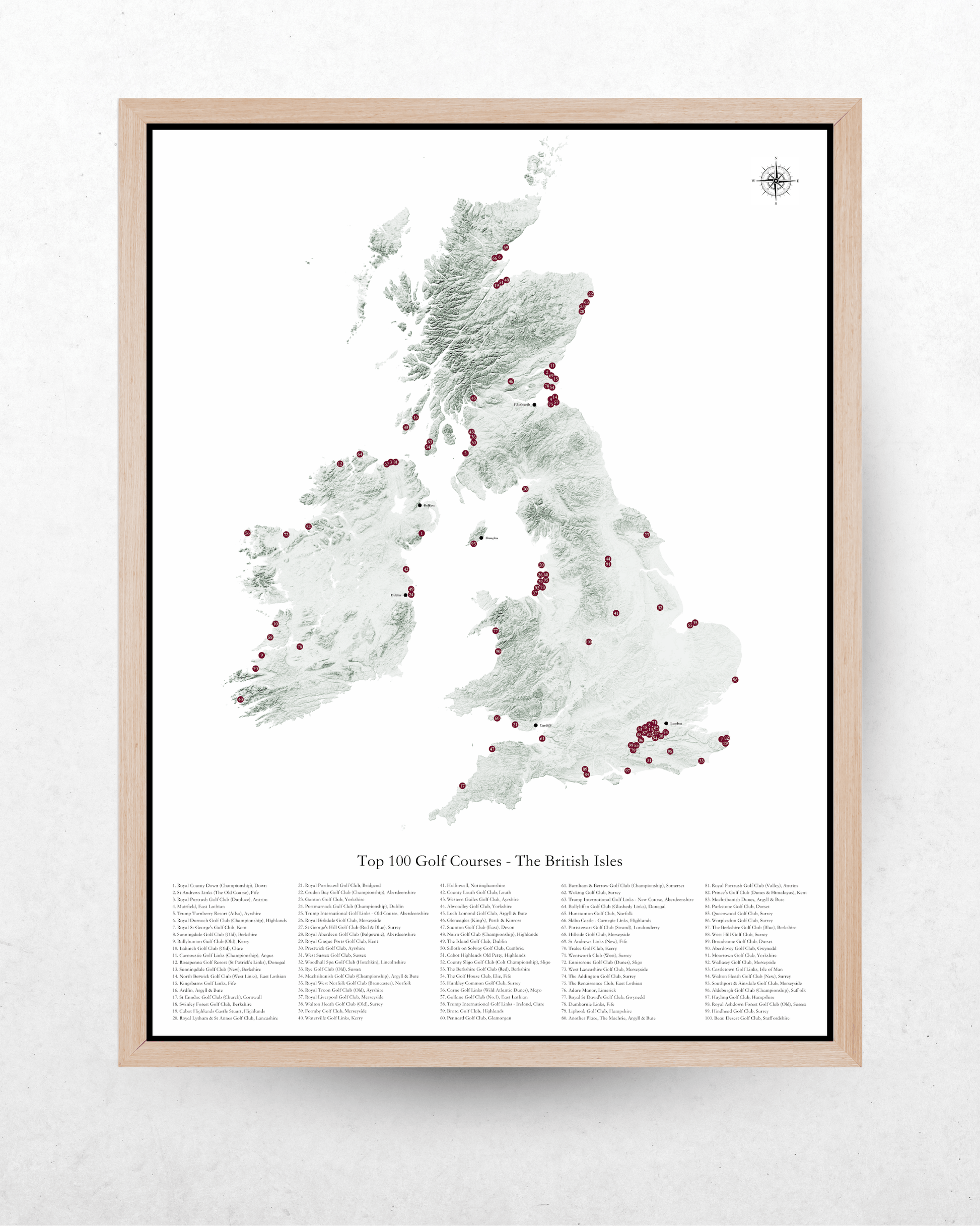 A framed map print of the British Isles highlighting the top 100 golf courses, featuring a light wood frame against a textured white wall.